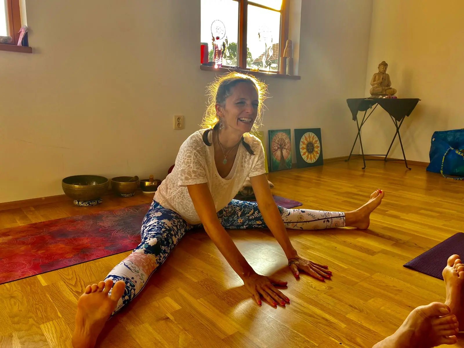 Žena sedí so skríženými nohami na farebnej podložke a smeje sa, v pozadí presvetlená miestnosť.