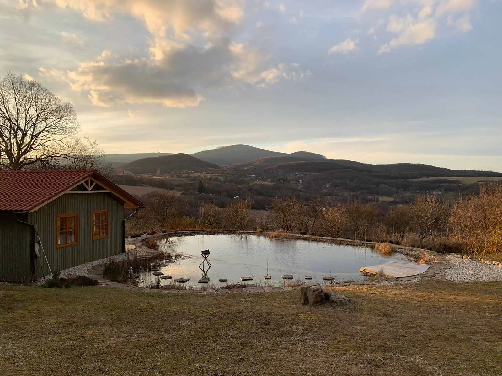 Dom pri jazere v podvečernom svetle, na hladine sa odrážajú kopce a oblaky.
